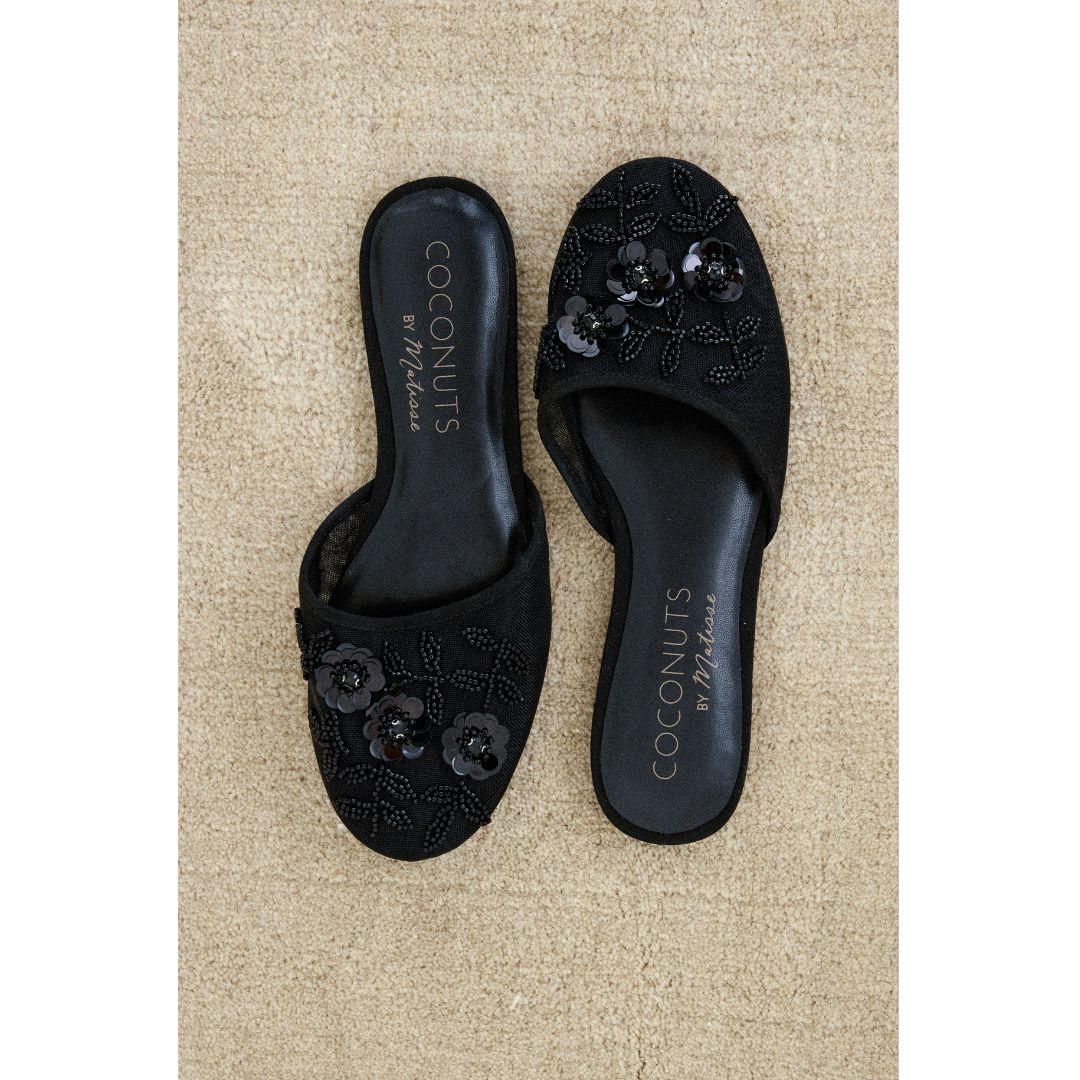 Black embroidered slippers with 'Coconuts by Matisse' branding on a beige carpet.