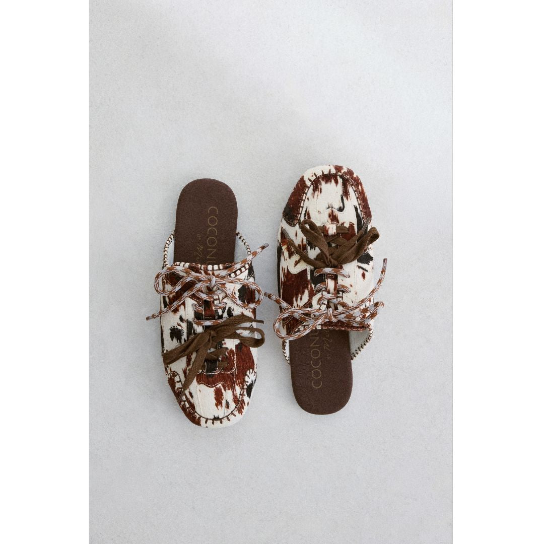 Pair of brown and white cow hair slip-on shoes with brown laces on a light gray background.