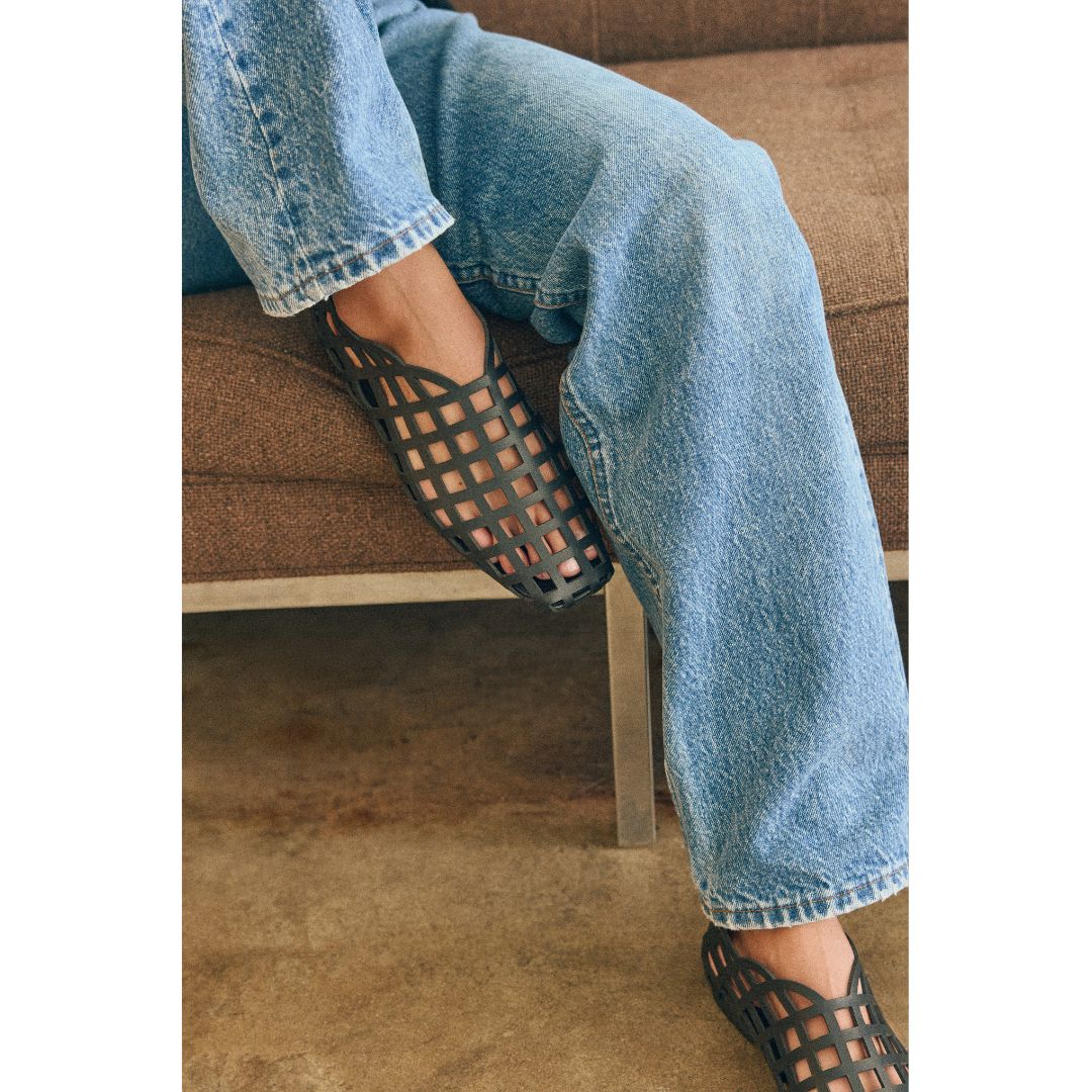 Person wearing blue jeans and black cage-style flat shoes sitting on a brown couch.