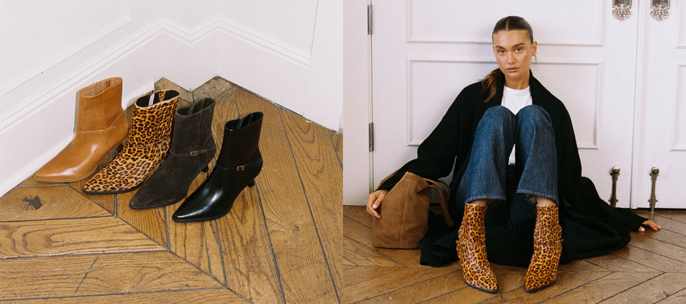 Collection of boots on a wooden floor with a person sitting against a white wall.