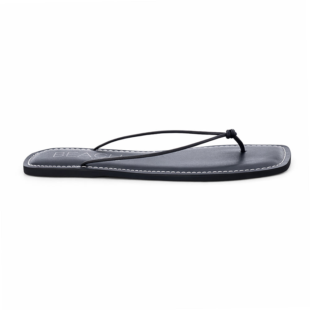 Black leather flat thong sandal with slim knotted straps and square toe, shown on a white background.