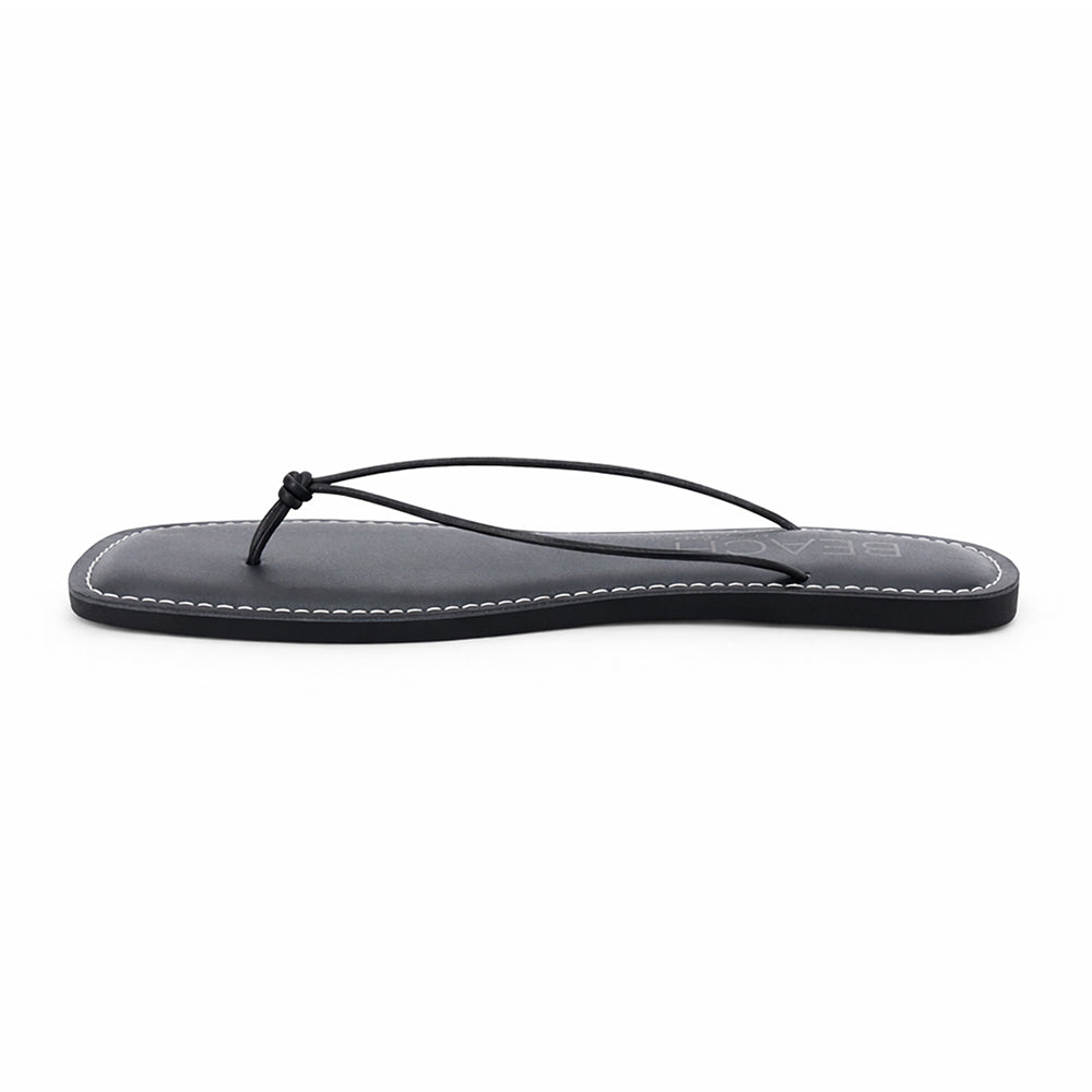 Black leather flat thong sandal with slim knotted straps and square toe, shown on a white background.