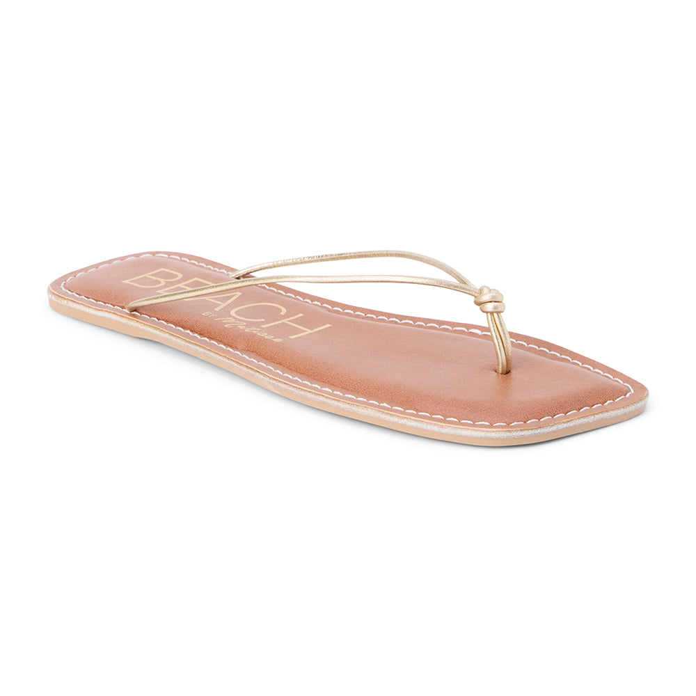 Metallic gold leather flat thong sandal with slim knotted straps and square toe, shown on a white background.