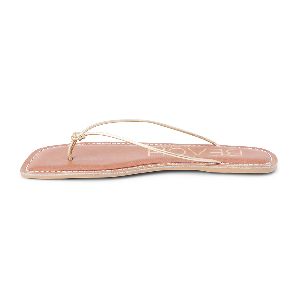 Metallic gold leather flat thong sandal with slim knotted straps and square toe, shown on a white background.