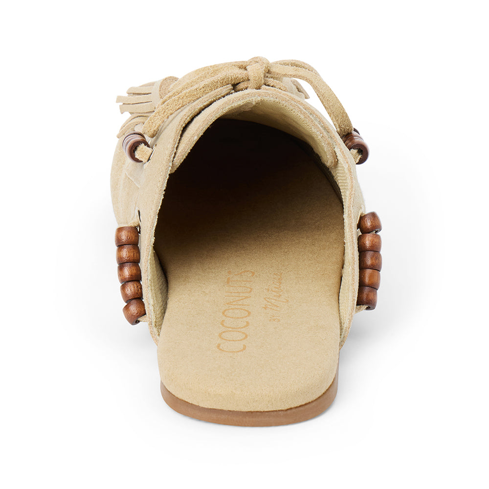 Tan suede mule with an open back, moc-toe stitching, lace-up tassels, wood bead accents, and a flat sole shown on a white background.
