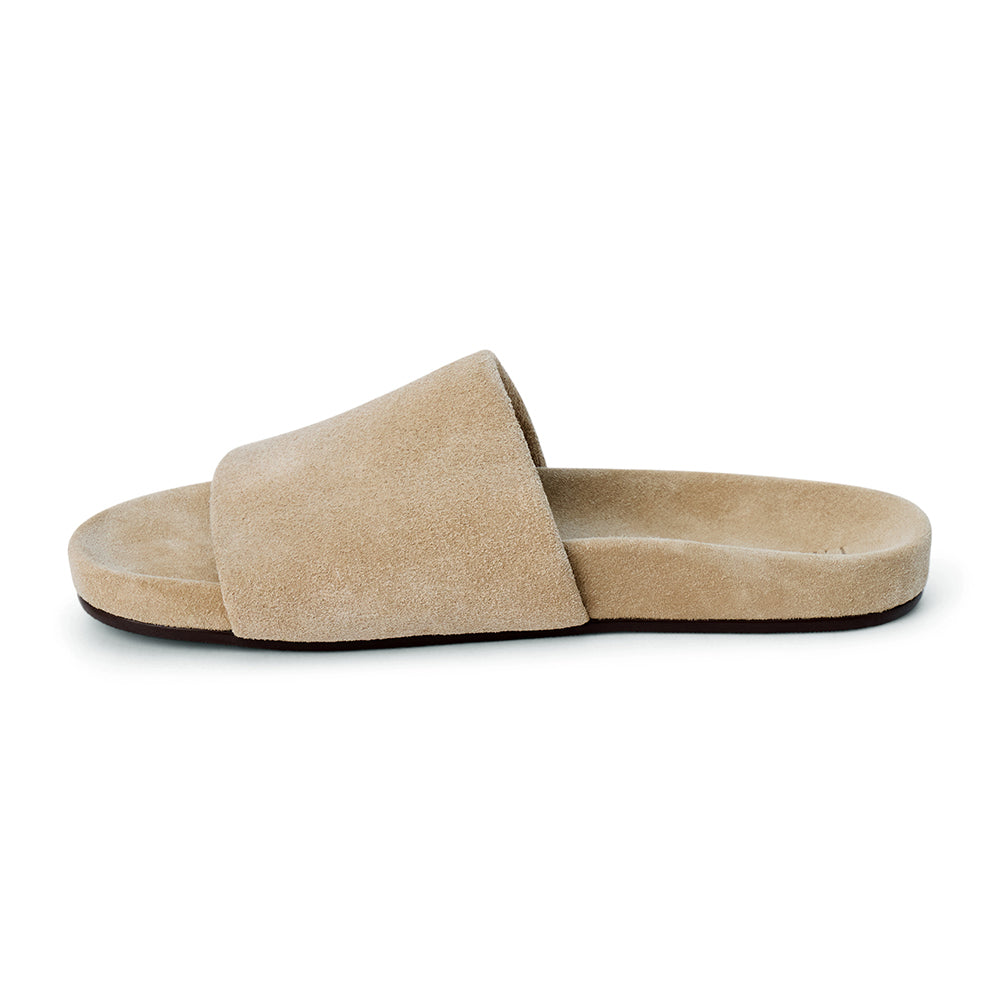 Sand beige suede flat slide sandal with wide strap and molded footbed, shown on a white background.