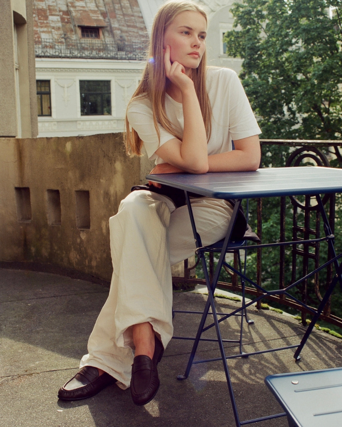 A woman sits at a blue folding table on an outdoor balcony, wearing a white short-sleeve T-shirt, cream wide-leg trousers, and dark loafers.