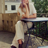 A woman sits at a blue folding table on an outdoor balcony, wearing a white short-sleeve T-shirt, cream wide-leg trousers, and dark loafers.