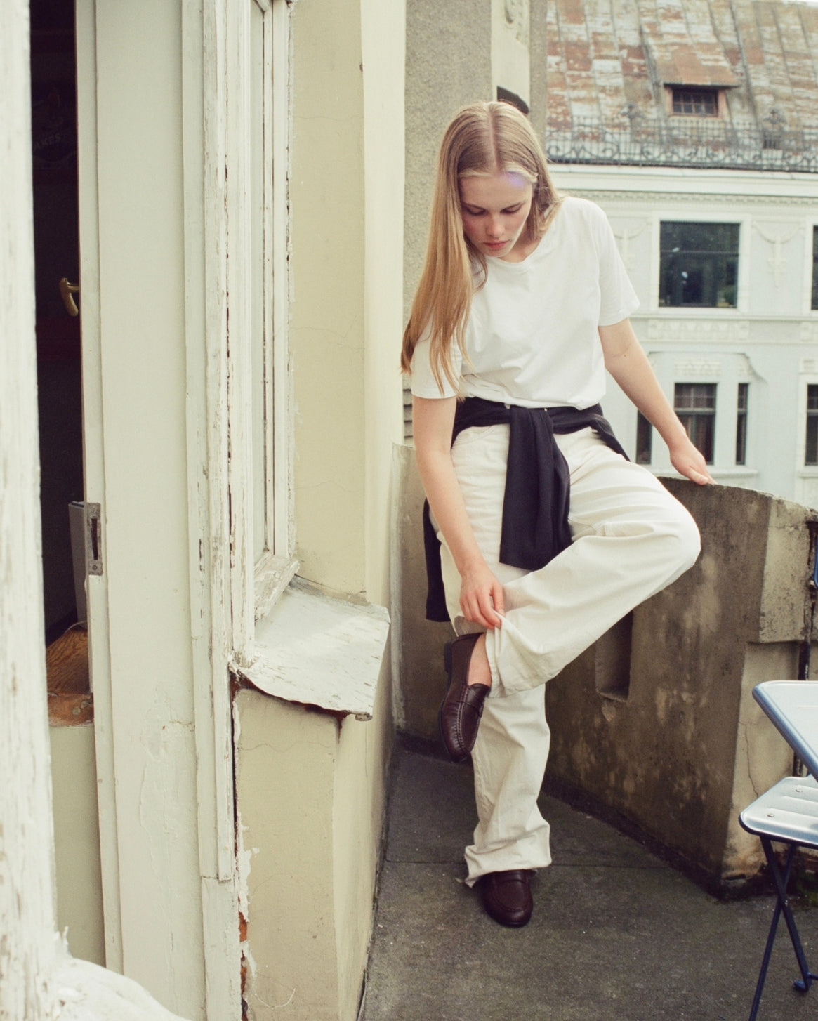 A woman stands on a balcony wearing a white short-sleeve t-shirt, white wide-leg trousers, and dark brown leather shoes, with a black shirt tied around her waist.