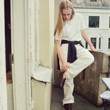 A woman stands on a balcony wearing a white short-sleeve t-shirt, white wide-leg trousers, and dark brown leather shoes, with a black shirt tied around her waist.
