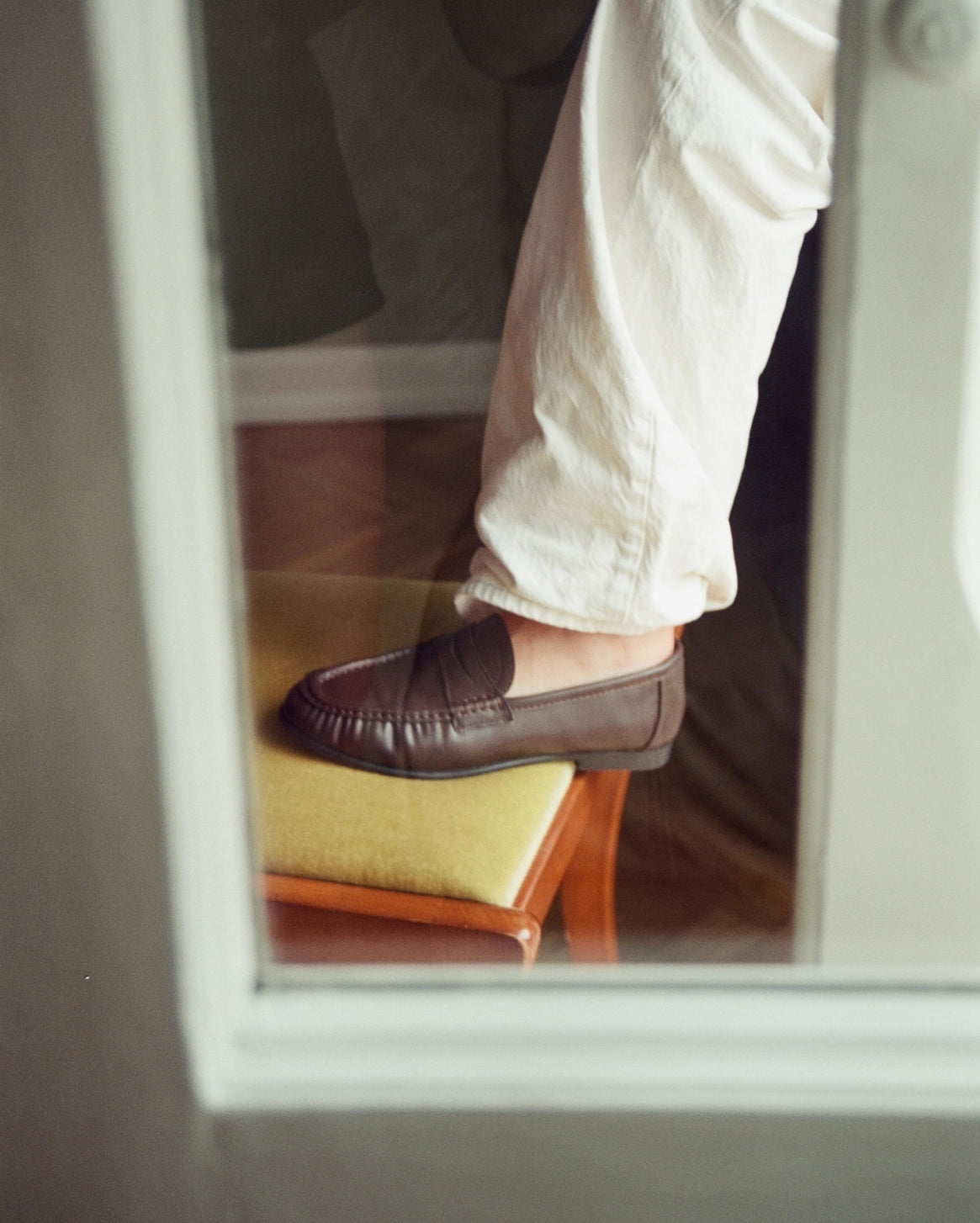 A person wearing off-white trousers and a dark brown leather penny loafer sits in a chair, viewed through a window or mirror reflection.