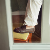 A person wearing off-white trousers and a dark brown leather penny loafer sits in a chair, viewed through a window or mirror reflection.