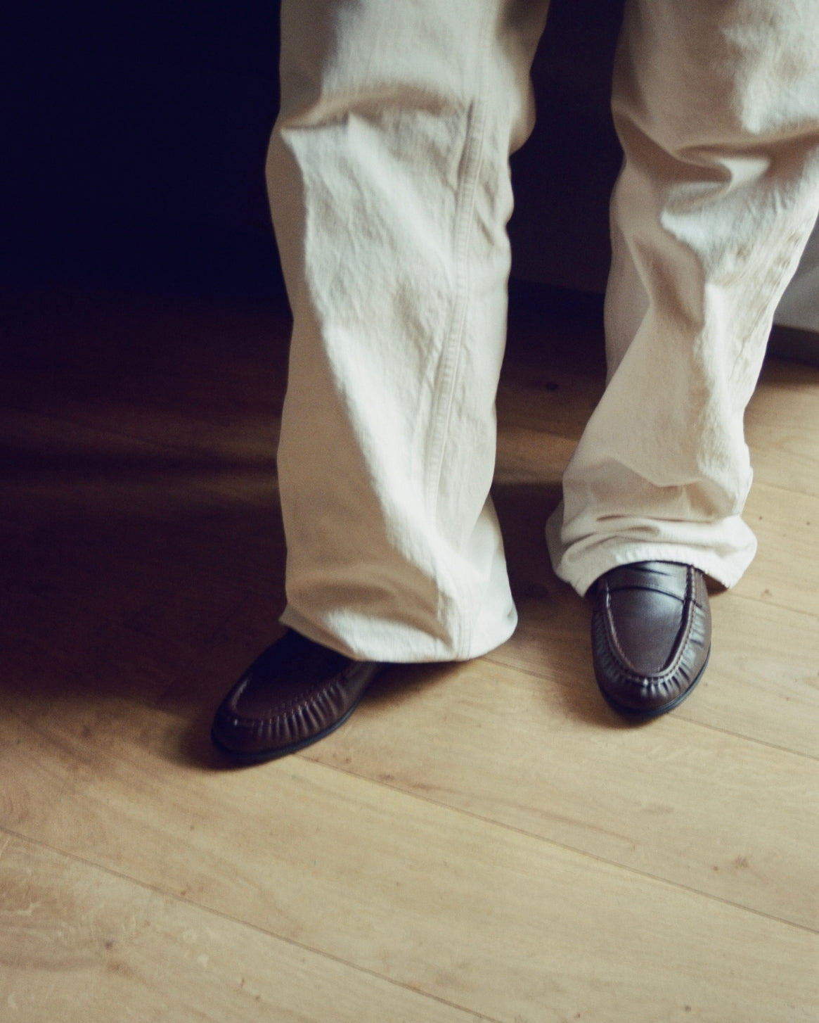 A person wearing off-white linen trousers and dark brown leather moccasin-style shoes, standing on a light wood floor.