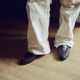 A person wearing off-white linen trousers and dark brown leather moccasin-style shoes, standing on a light wood floor.