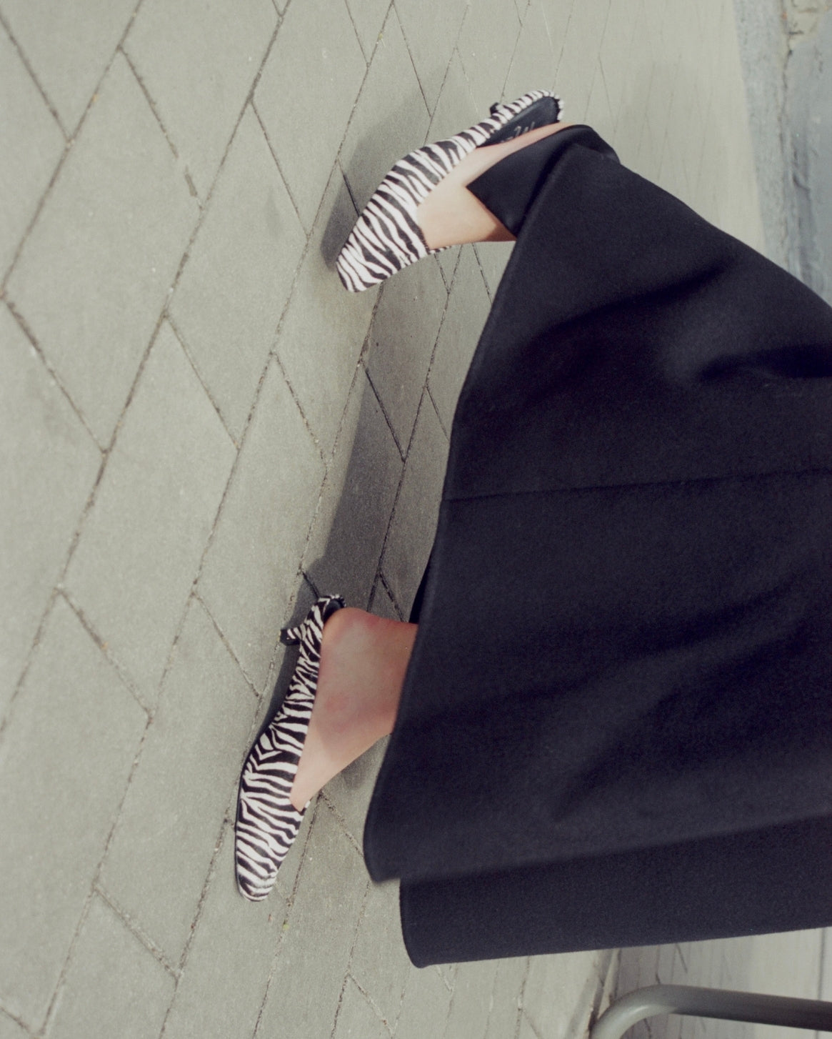 A person wearing black pointed flats with a zebra print pattern, paired with a black skirt, standing on a gray paved surface.