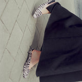 A person wearing black pointed flats with a zebra print pattern, paired with a black skirt, standing on a gray paved surface.