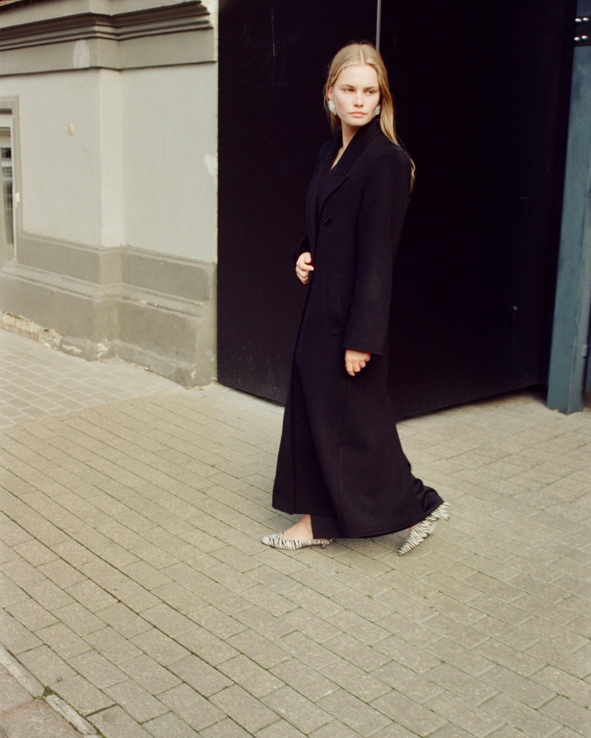 A woman wearing a long black coat and patterned flat shoes walks on a paved street next to a building with a dark doorway.