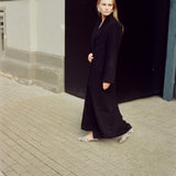 A woman wearing a long black coat and patterned flat shoes walks on a paved street next to a building with a dark doorway.