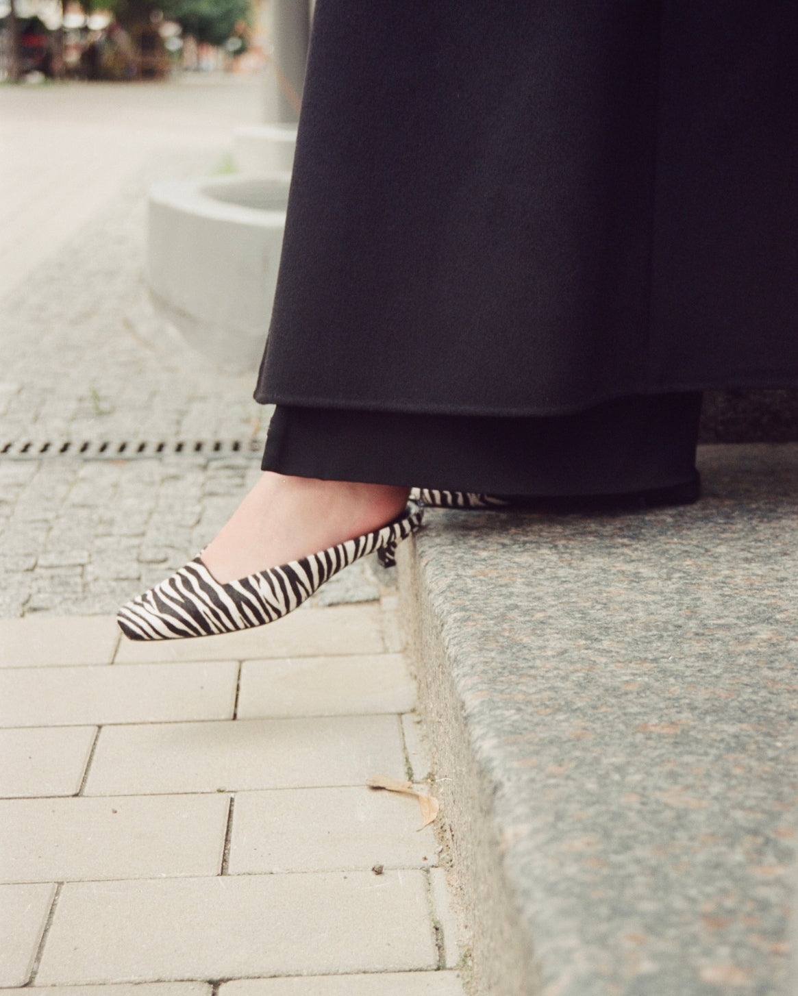 A person wearing black wide-leg trousers and black-and-white zebra-pattern pointed-toe slingback flats, standing on a stone curb outdoors.