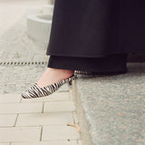 A person wearing black wide-leg trousers and black-and-white zebra-pattern pointed-toe slingback flats, standing on a stone curb outdoors.
