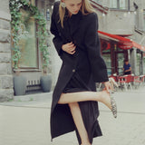 A woman in a long black coat and black skirt stands on a city sidewalk, holding up one leg to show black-and-white zebra-patterned pointed-toe flats. Buildings, windows, and outdoor seating are visible in the background.