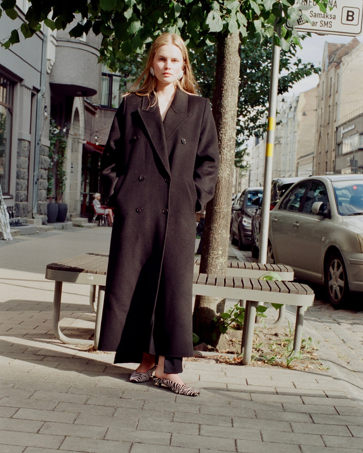 A woman stands on a city sidewalk wearing a long black double-breasted coat over a white top and leopard-print flat shoes, with trees, benches, parked cars, and buildings in the background.