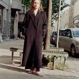 A woman stands on a city sidewalk wearing a long black double-breasted coat over a white top and leopard-print flat shoes, with trees, benches, parked cars, and buildings in the background.