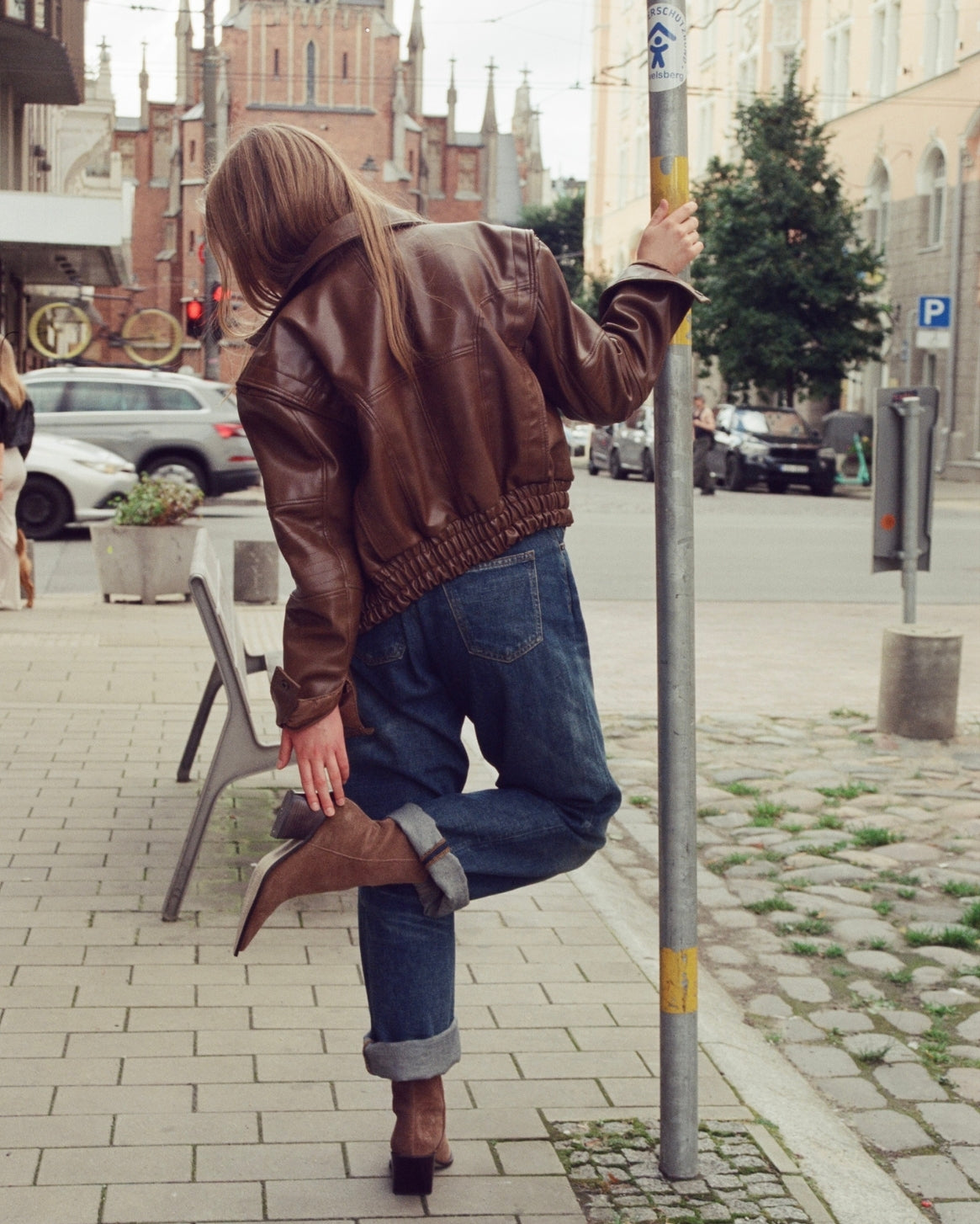 A woman wearing a brown leather bomber jacket, blue jeans, and brown ankle boots is photographed from behind in an outdoor city street setting.