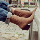 A person wearing brown suede pointed-toe ankle boots with a block heel, paired with cuffed blue jeans, sitting on a metal bench in an outdoor urban setting.