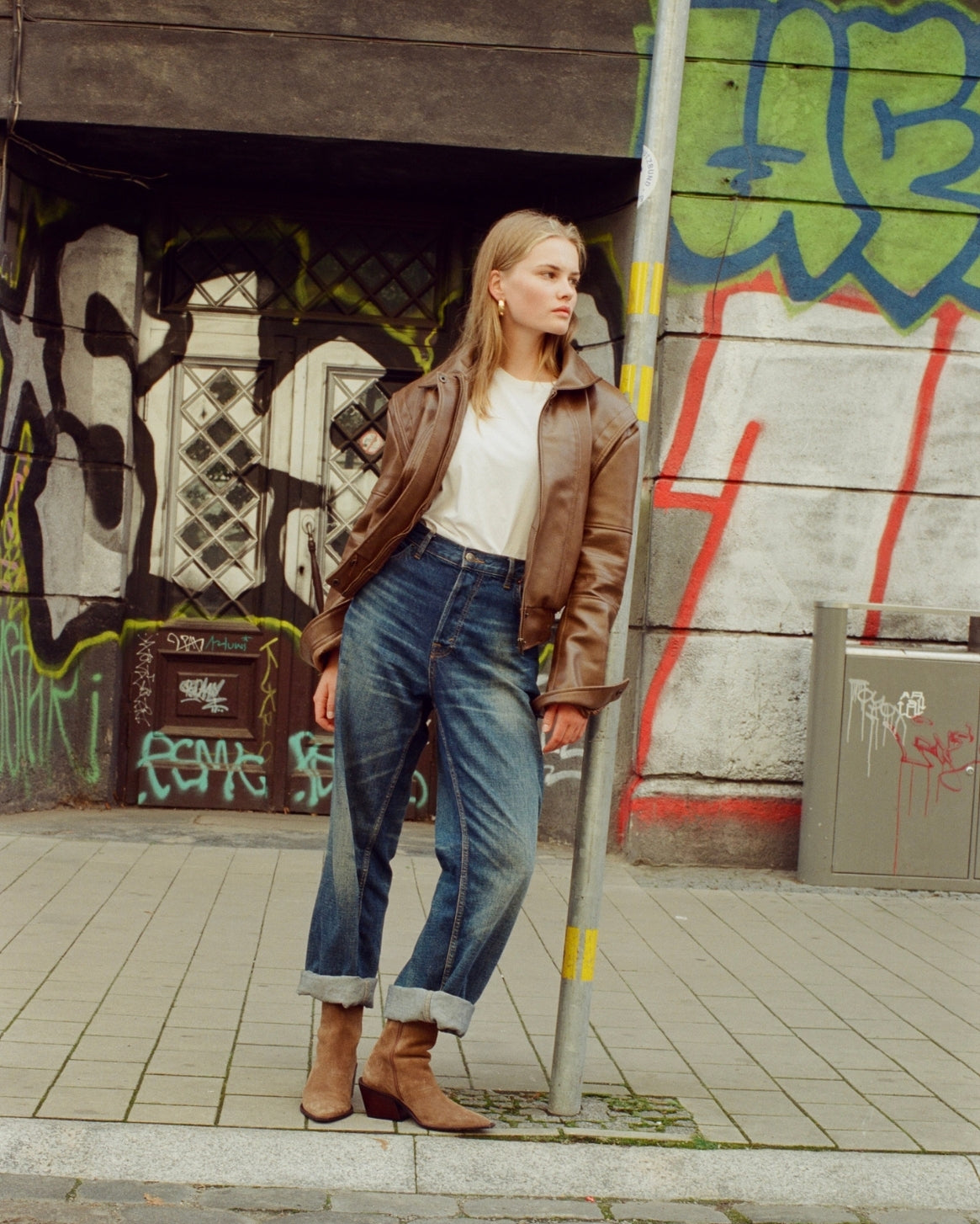 A woman stands on a city sidewalk in front of a graffiti-covered wall, wearing a brown leather jacket, white top, blue jeans, and brown ankle boots.