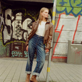 A woman stands on a city sidewalk in front of a graffiti-covered wall, wearing a brown leather jacket, white top, blue jeans, and brown ankle boots.