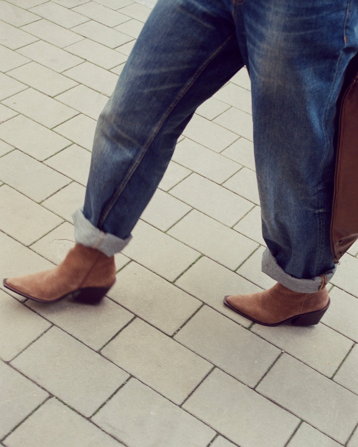 A person wearing blue jeans with rolled cuffs and brown pointed-toe ankle boots, walking on a light gray paved surface.