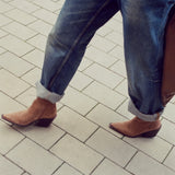 A person wearing blue jeans with rolled cuffs and brown pointed-toe ankle boots, walking on a light gray paved surface.