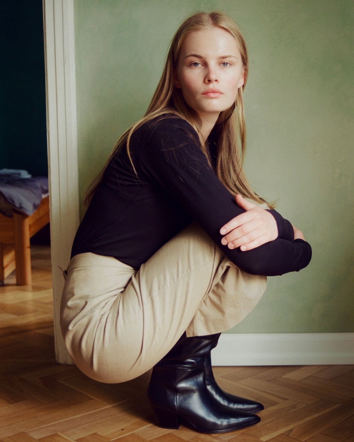 A woman crouching in a room with a green wall and wood floor, wearing a black long-sleeve top, beige pants, and black boots.
