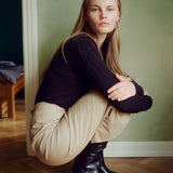 A woman crouching in a room with a green wall and wood floor, wearing a black long-sleeve top, beige pants, and black boots.