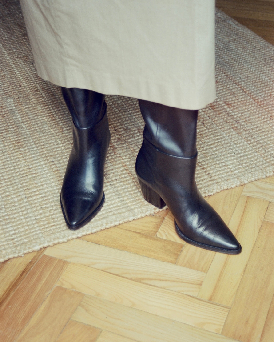 Black leather pointed-toe boots with low block heels, shown on a model wearing a beige skirt, standing on a woven jute rug over a light wood parquet floor.