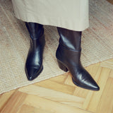 Black leather pointed-toe boots with low block heels, shown on a model wearing a beige skirt, standing on a woven jute rug over a light wood parquet floor.