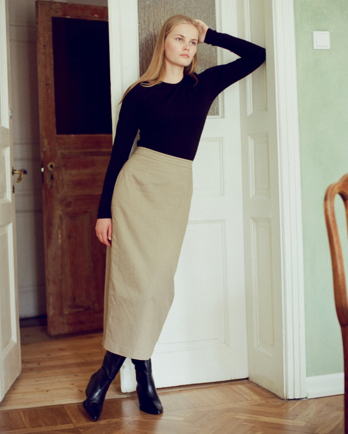 A woman stands in a doorway wearing a black long-sleeve top and a beige midi skirt, paired with black boots.
