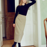 A woman stands in a doorway wearing a black long-sleeve top and a beige midi skirt, paired with black boots.