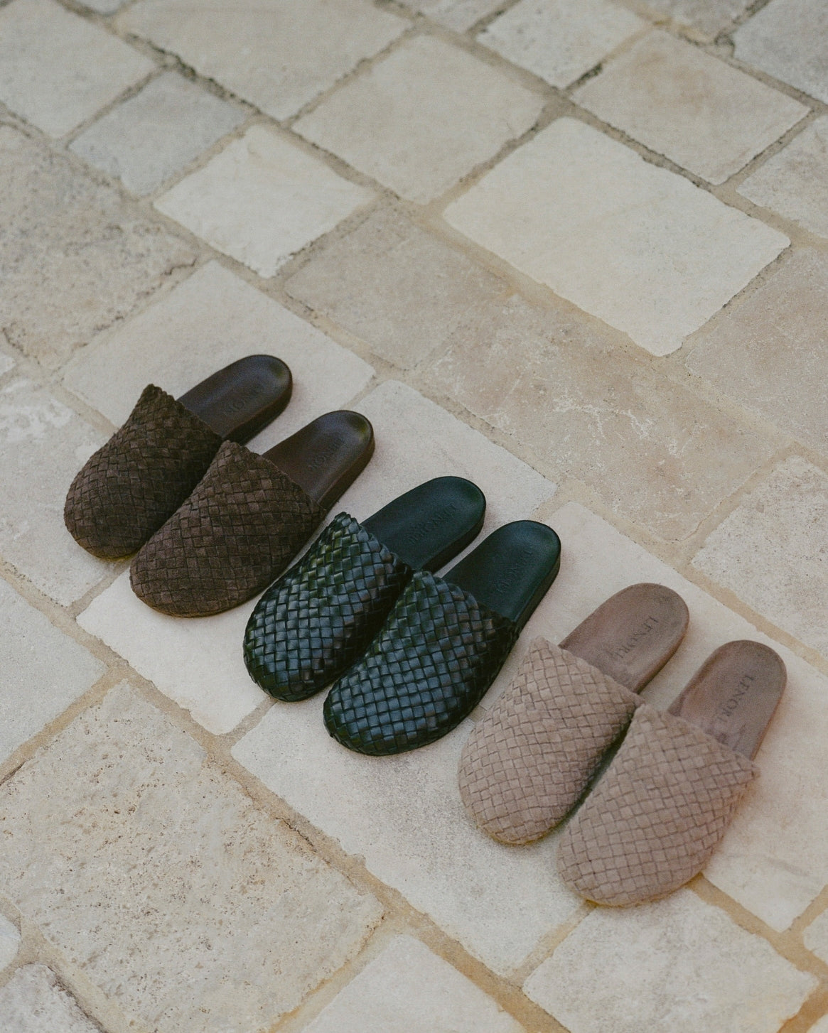 Four woven slip-on mules arranged diagonally on a light beige stone tile surface. The slippers come in dark brown, dark green, and light beige/tan woven leather.
