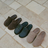 Four woven slip-on mules arranged diagonally on a light beige stone tile surface. The slippers come in dark brown, dark green, and light beige/tan woven leather.
