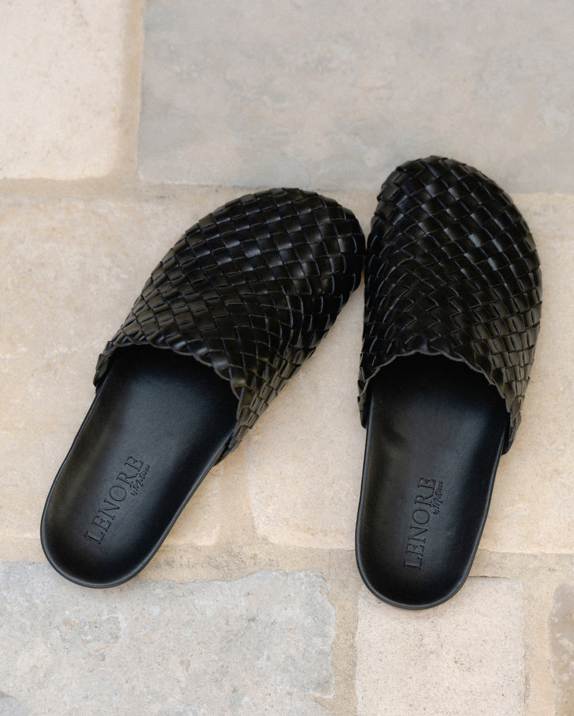 A pair of black woven leather mules with scalloped edges, displayed on a light beige stone tile surface. The brand name 'LENORE' is debossed on the insoles.