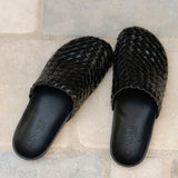A pair of black woven leather mules with scalloped edges, displayed on a light beige stone tile surface. The brand name 'LENORE' is debossed on the insoles.