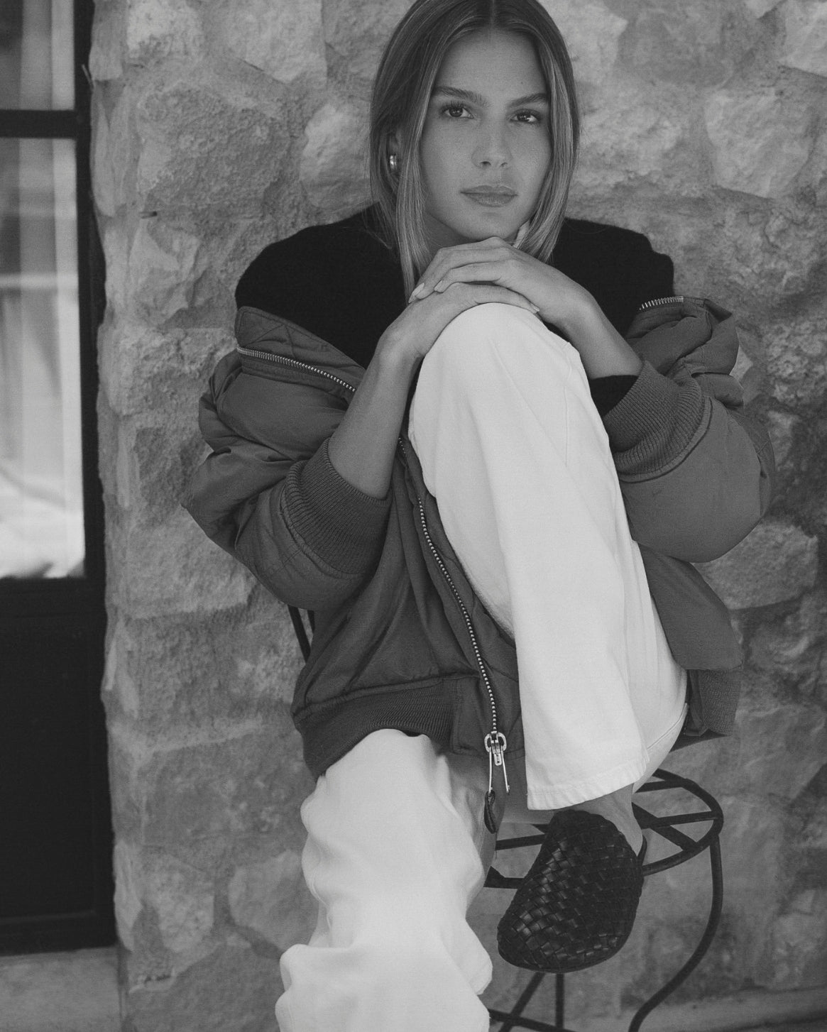 A black and white lifestyle photograph of a woman sitting on a metal stool against a rustic stone wall. She is wearing a dark oversized zip-up jacket, light-colored wide-leg pants, and a dark woven slipper.