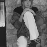 A black and white lifestyle photograph of a woman sitting on a metal stool against a rustic stone wall. She is wearing a dark oversized zip-up jacket, light-colored wide-leg pants, and a dark woven slipper.