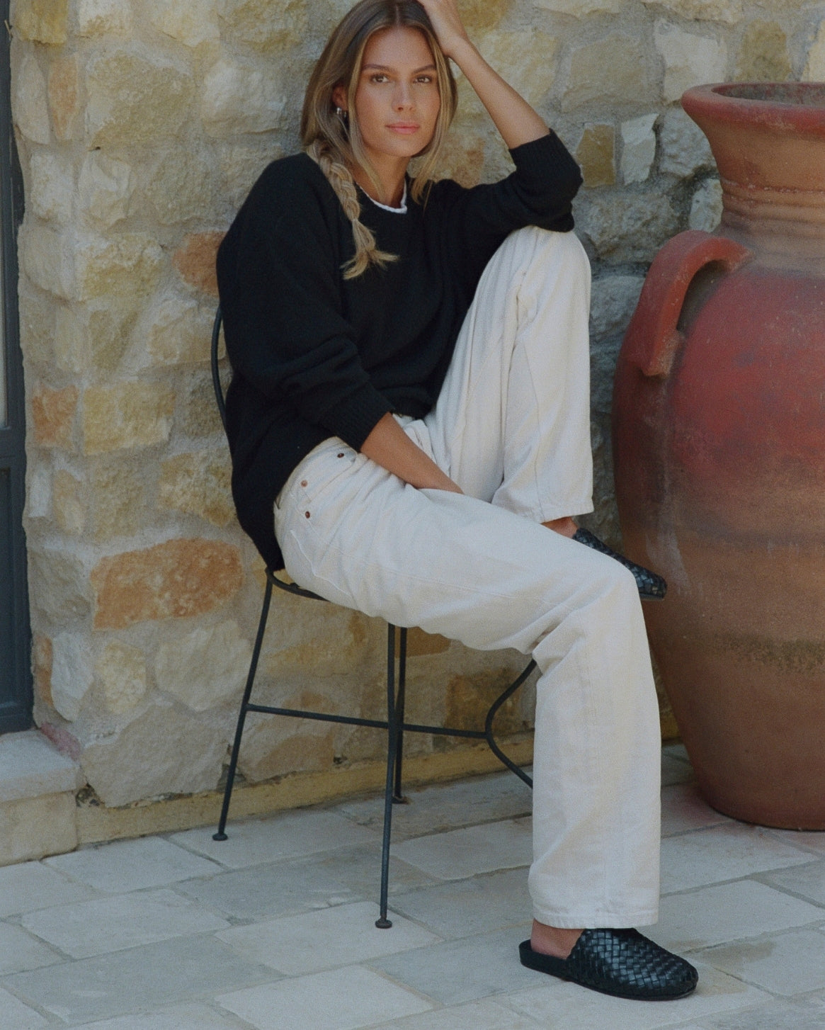 A woman sits on a black metal chair outdoors against a stone wall, wearing a black long-sleeve top, off-white wide-leg trousers, and black woven slip-on shoes. A large terracotta pot is visible to her right.