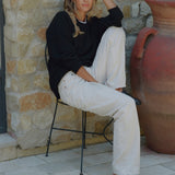 A woman sits on a black metal chair outdoors against a stone wall, wearing a black long-sleeve top, off-white wide-leg trousers, and black woven slip-on shoes. A large terracotta pot is visible to her right.
