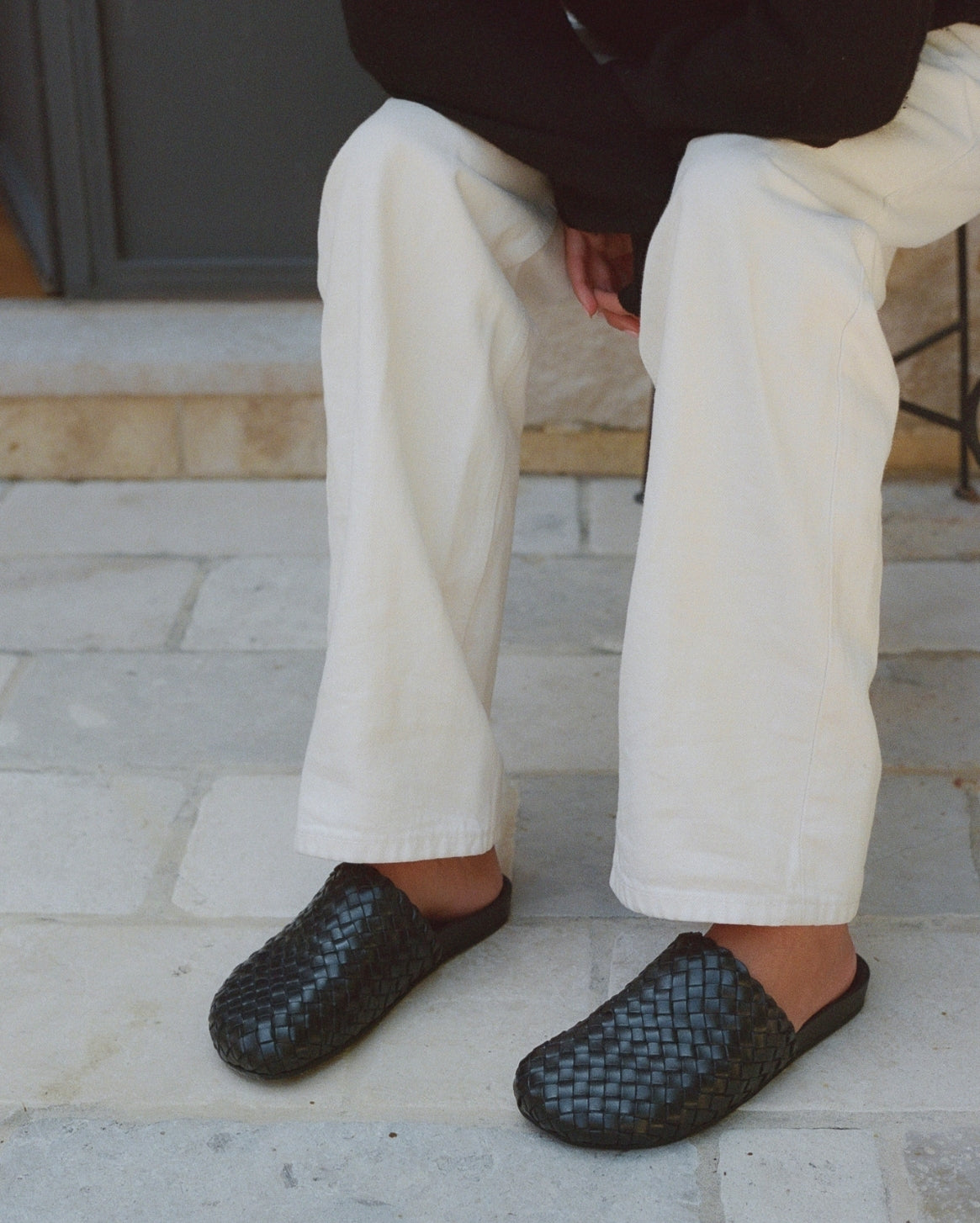 A person seated outdoors wearing off-white wide-leg trousers and dark woven slip-on shoes on a light stone patio.