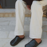 A person seated outdoors wearing off-white wide-leg trousers and dark woven slip-on shoes on a light stone patio.