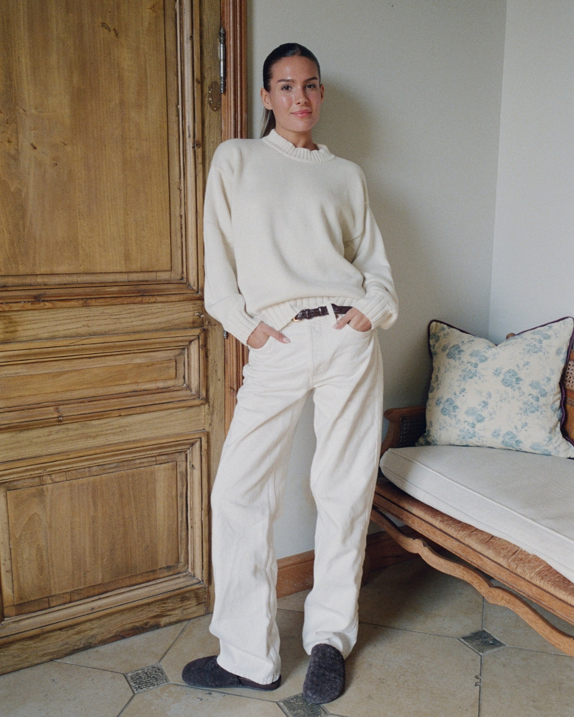 Person wearing a white sweater and pants standing in a room with wooden door and floral-patterned cushion.